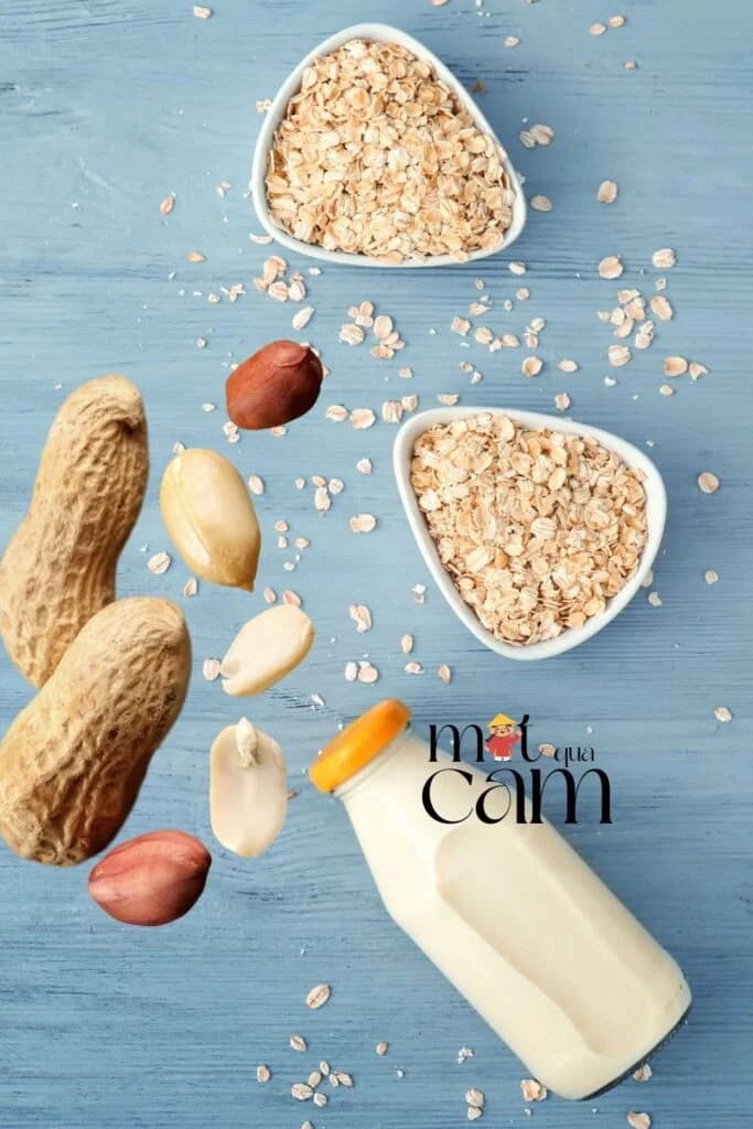 Chai sữa yến mạch đậu phộng cùng hai bát yến mạch và lạc còn vỏ trên nền gỗ xanh
