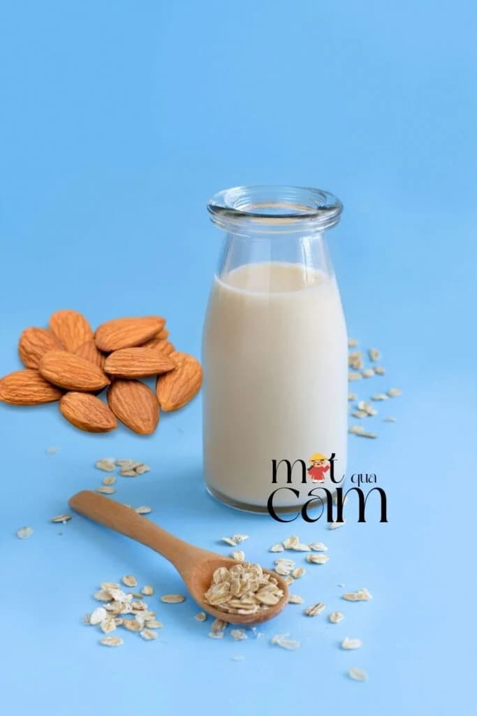 Chai sữa yến mạch hạnh nhân trên nền xanh, bên cạnh là hạnh nhân và thìa gỗ đựng yến mạch