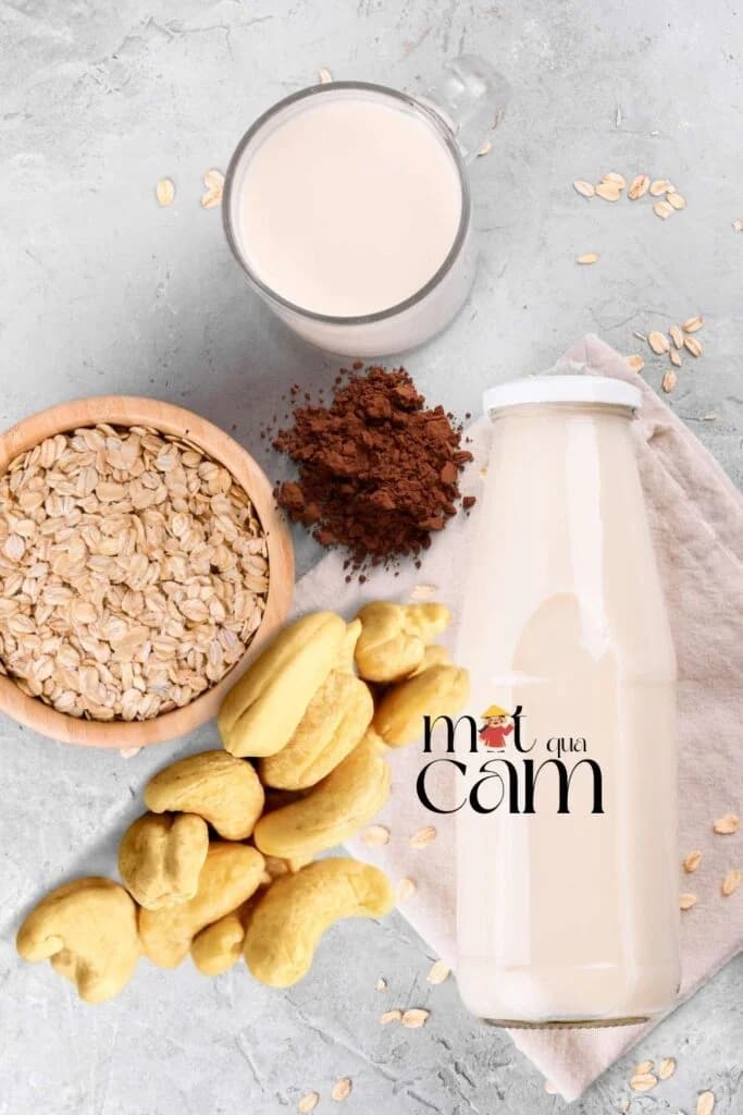 Chai và ly sữa yến mạch hạt điều cacao, bên cạnh là bát yến mạch, bột cacao và hạt điều